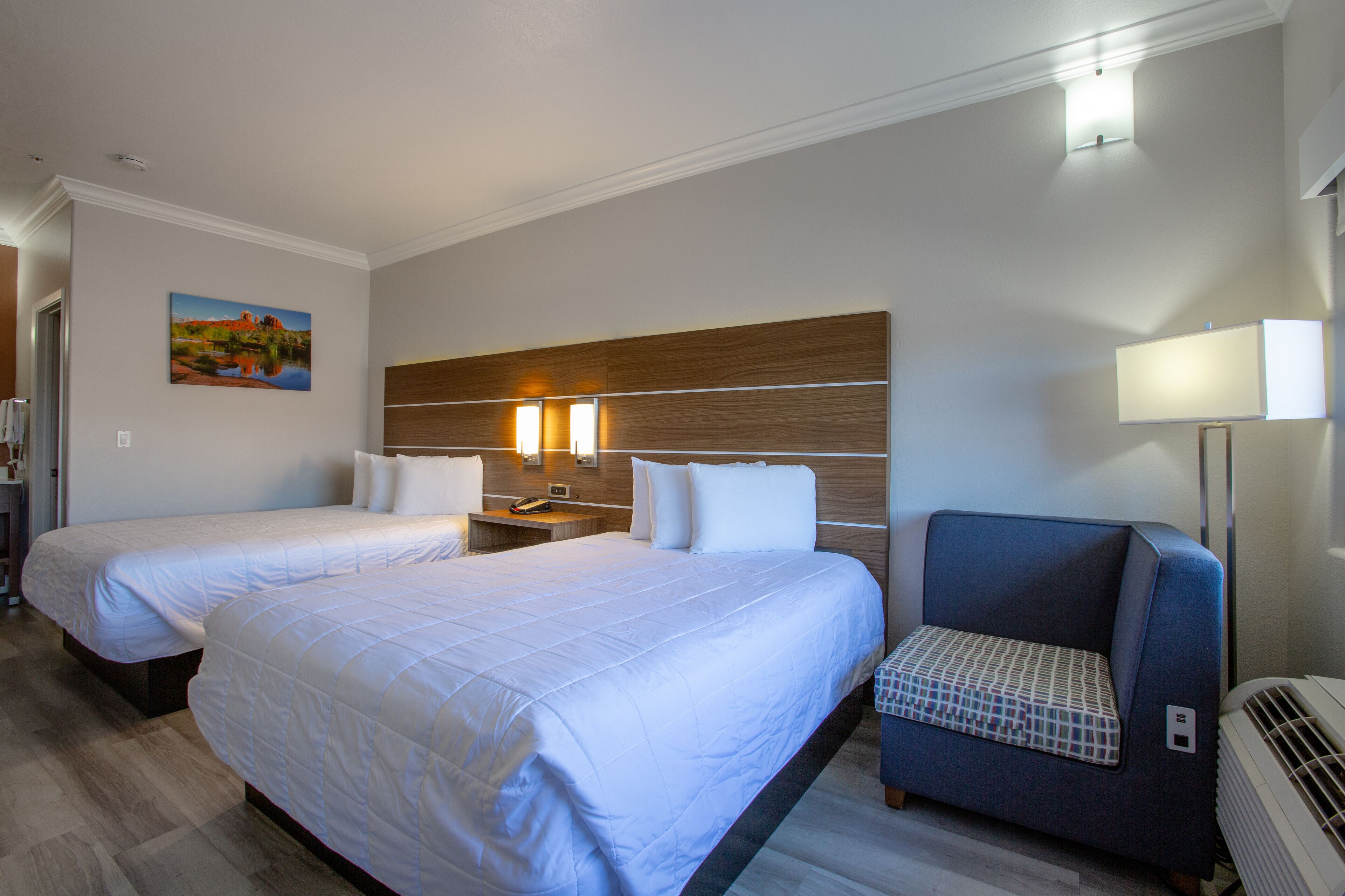 A hotel room with two beds, a nightstand with a lamp, a chair, and a painting on the wall.