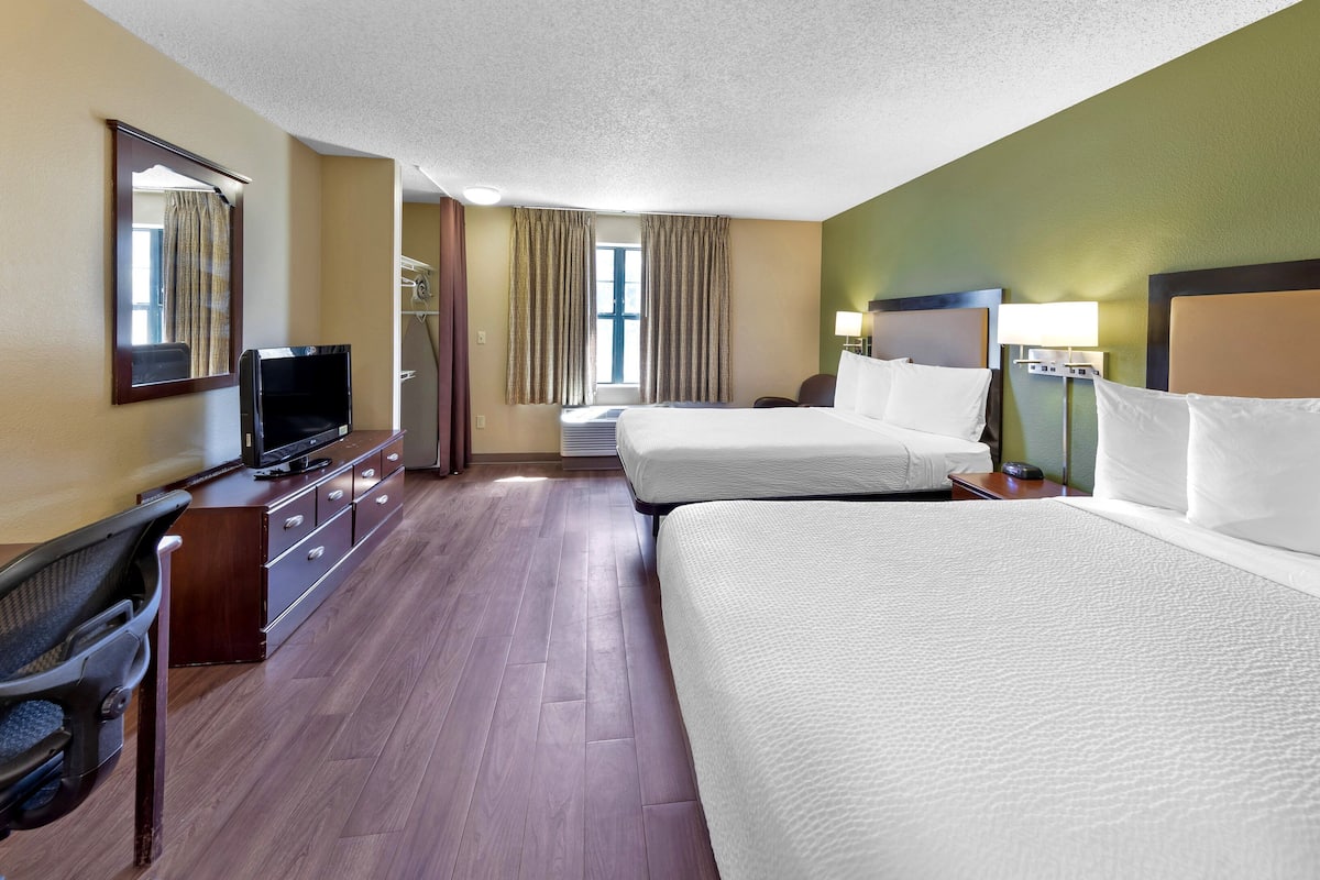 A hotel room with two beds, a desk with a television, a chair, and a window with curtains.