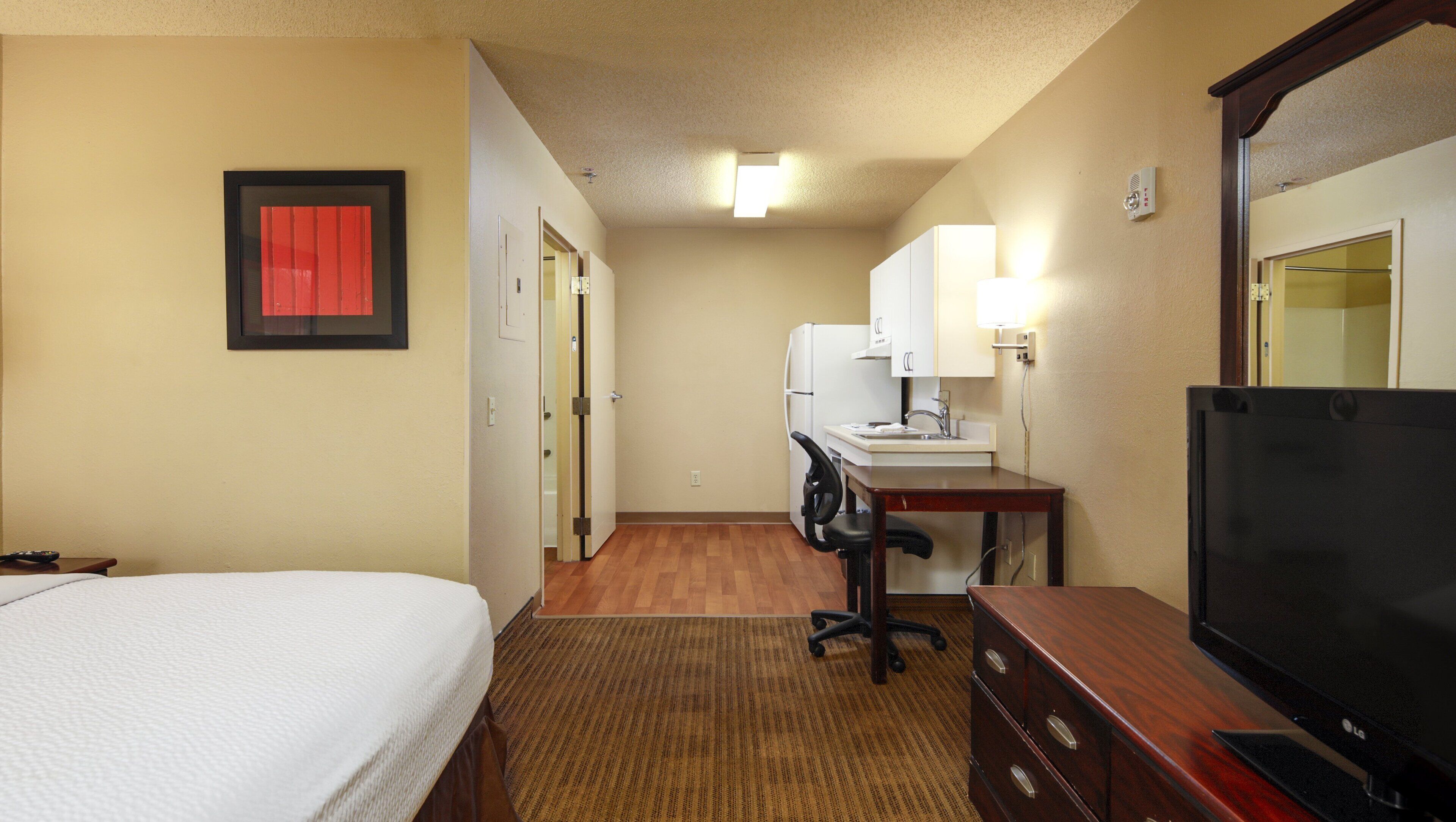 Une chambre d’hôtel comprenant un lit, un bureau avec une chaise, une télévision, un réfrigérateur et une salle de bain visible à travers une porte ouverte.