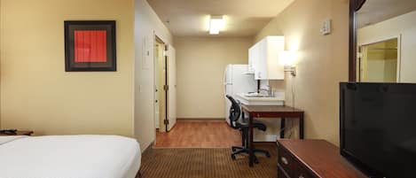 Quarto de hotel com cama, escrivaninha e cadeira, televisão, frigorífico e casa de banho visível através de uma porta aberta.
