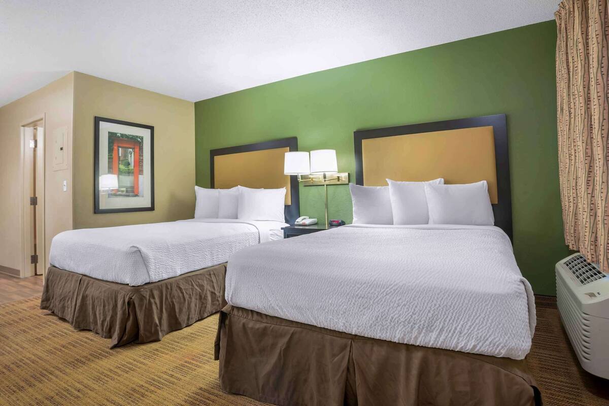 Camera d'albergo con due letti, una parete verde decorativa, un quadro incorniciato, un comodino con una lampada e una finestra con tende.
