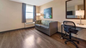Habitación de hotel con sofá, escritorio y silla, espejo grande, un cuadro y ventana con cortinas.