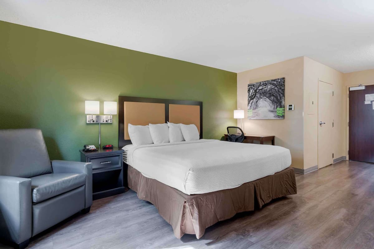 Habitación de hotel con cama, una silla, una mesita de noche, una lámpara y un cuadro en la pared.
