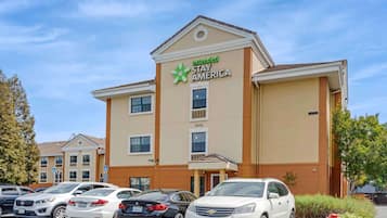 A Stay America hotel with a beige facade, multiple windows, and a parking lot with several cars.