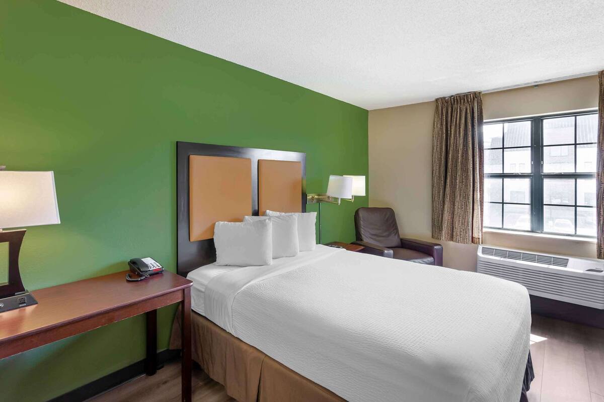Ein Hotelzimmer mit grüner Wand, einem Bett mit weißer Bettwäsche, einem Schreibtisch mit Telefon und einem Sessel.