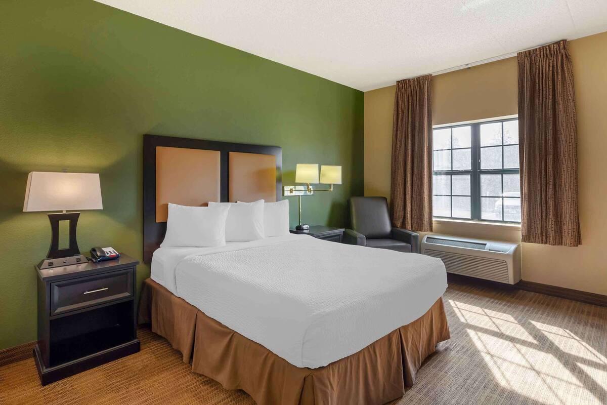 A hotel room with a large bed, a nightstand with a lamp, a chair, a window with curtains, and a wall-mounted heater.