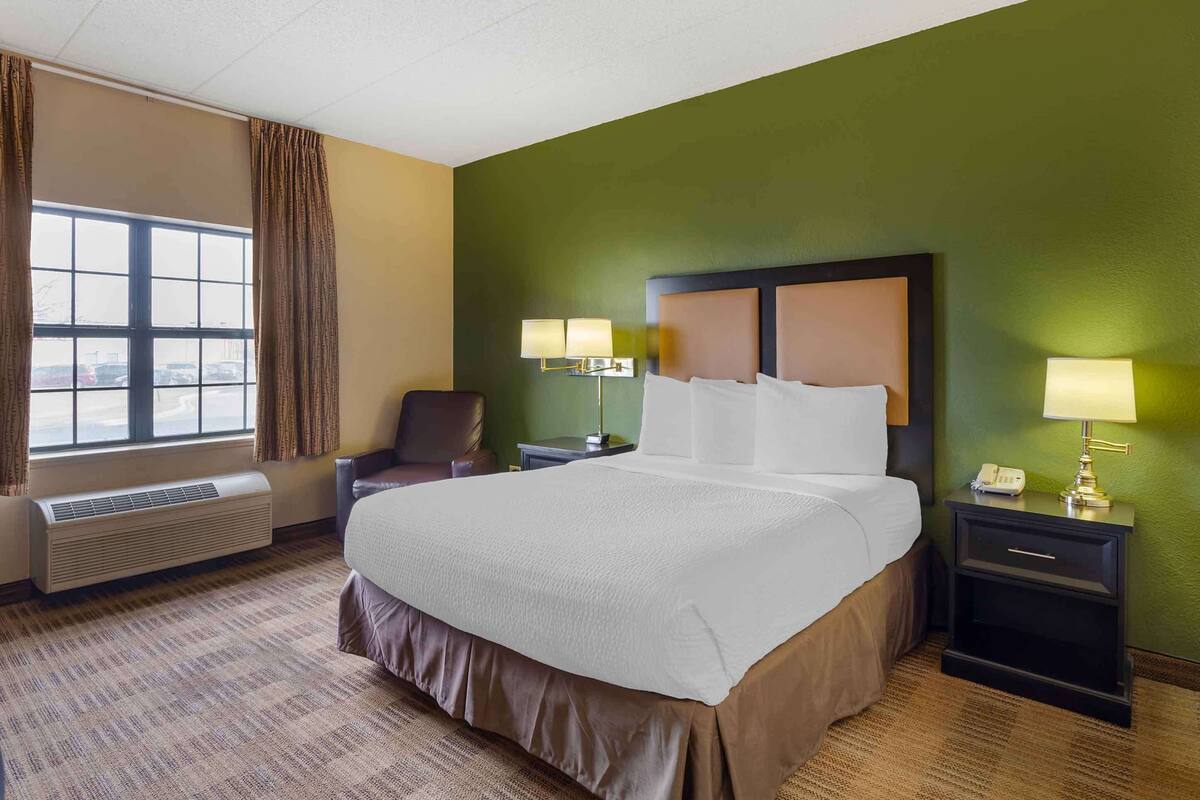 A hotel room with a large bed, a chair, a desk with a lamp, and a window with curtains.