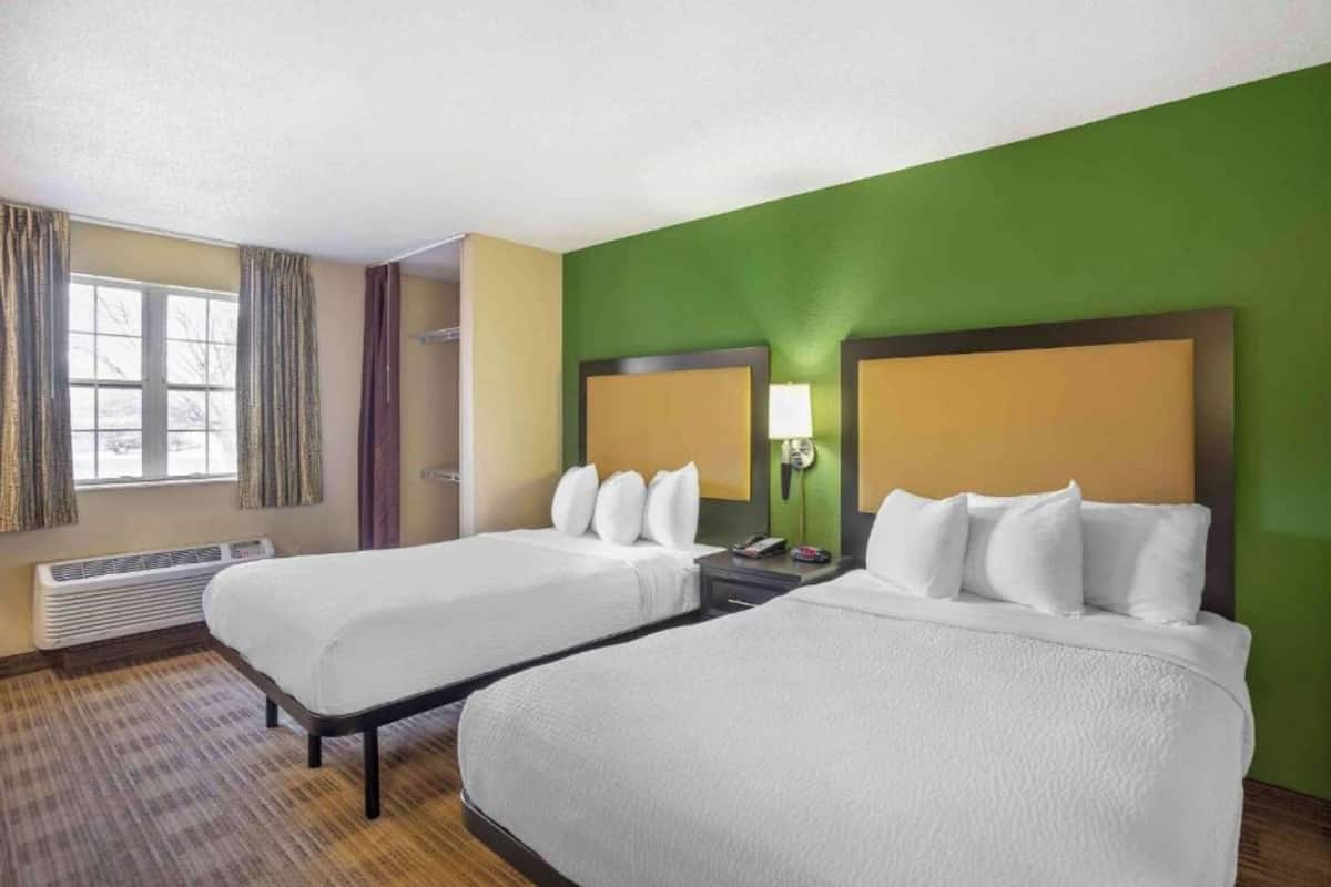 A hotel room with two beds, a window with curtains, a heater, and a green wall.