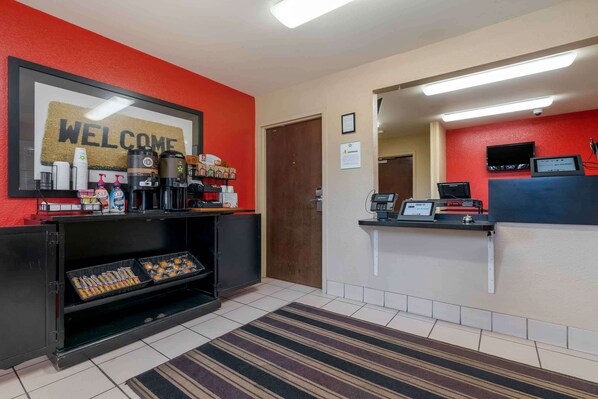 A reception area with a 'WELCOME' sign, coffee machines, a counter with a cash register, and a display of pastries.
