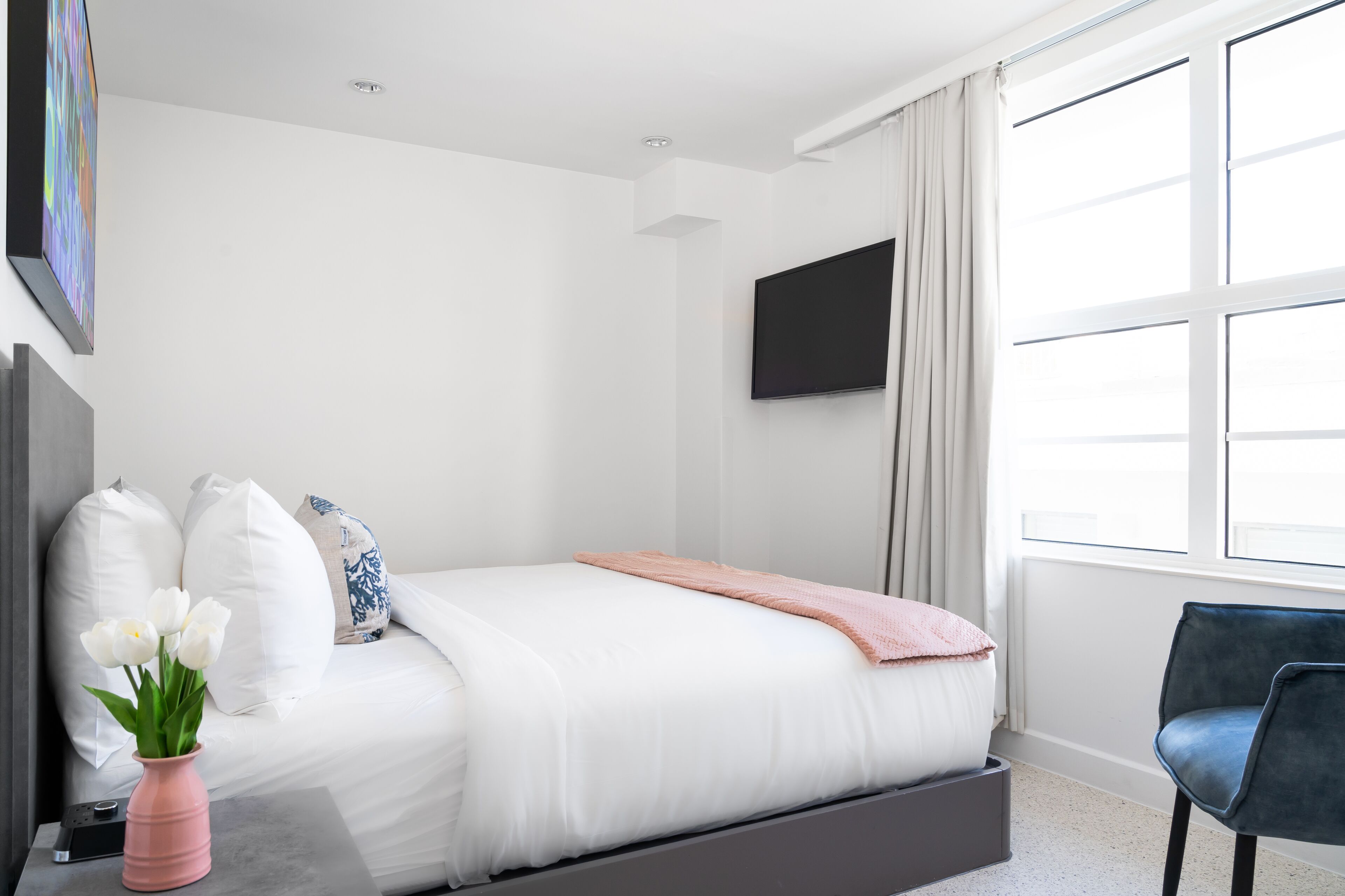 A modern bedroom with a bed, a flat-screen TV, a window with curtains, and a vase with flowers on a nightstand.