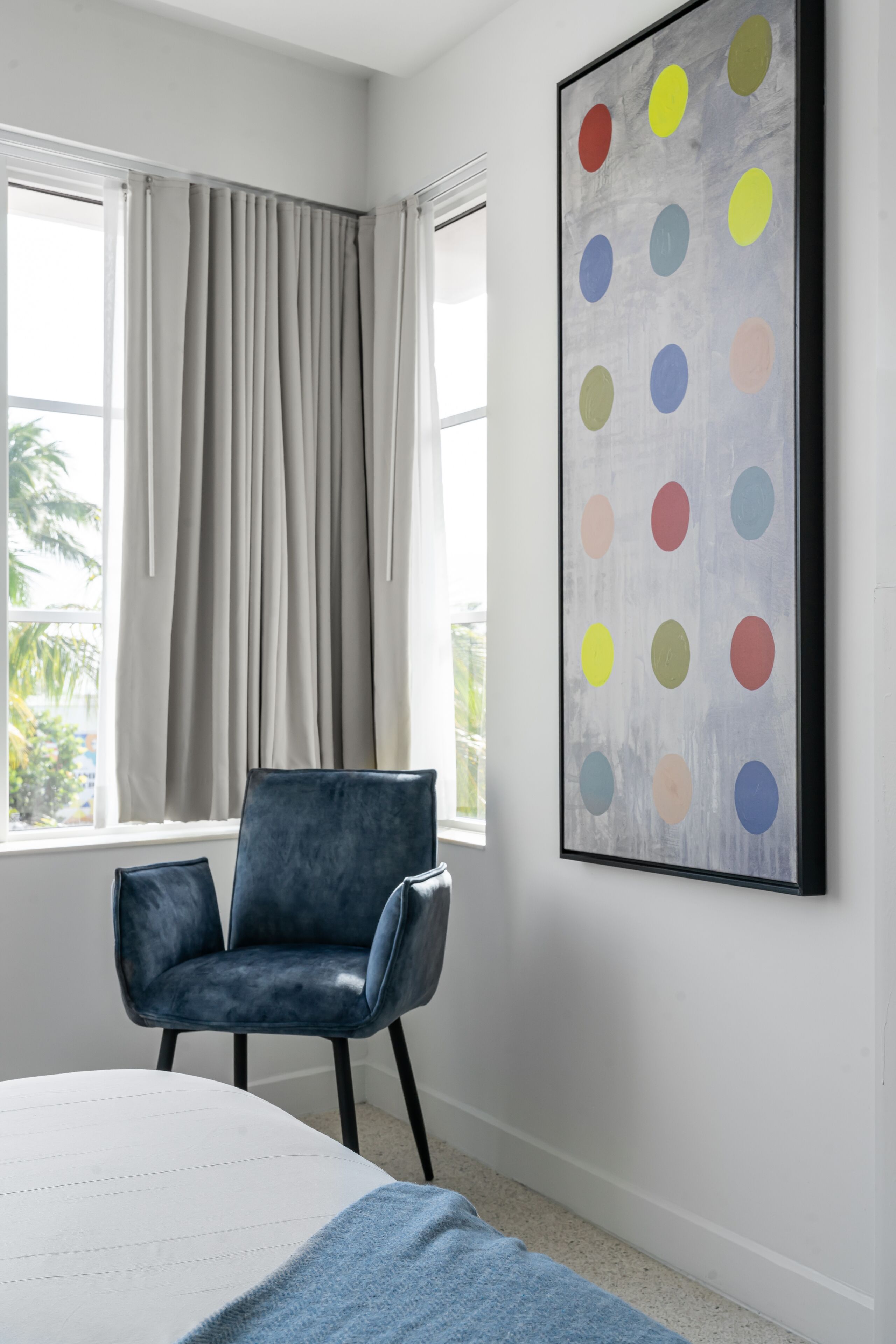 A bedroom with a bed, a chair, a window with curtains, and a colorful artwork on the wall.