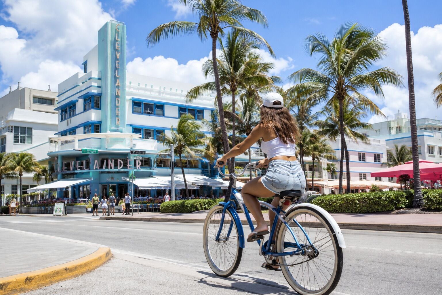 Una persona montando en bicicleta por una calle con palmeras y un edificio con letreros.