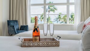 Ein Bett mit einer Champagnerflasche und zwei Gläsern auf einem Tablett, ein blauer Sessel und Blick auf Palmen durch ein Fenster.