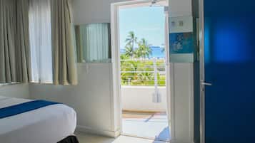 A hotel room with a bed, a door leading to a balcony with a view of palm trees and the ocean.
