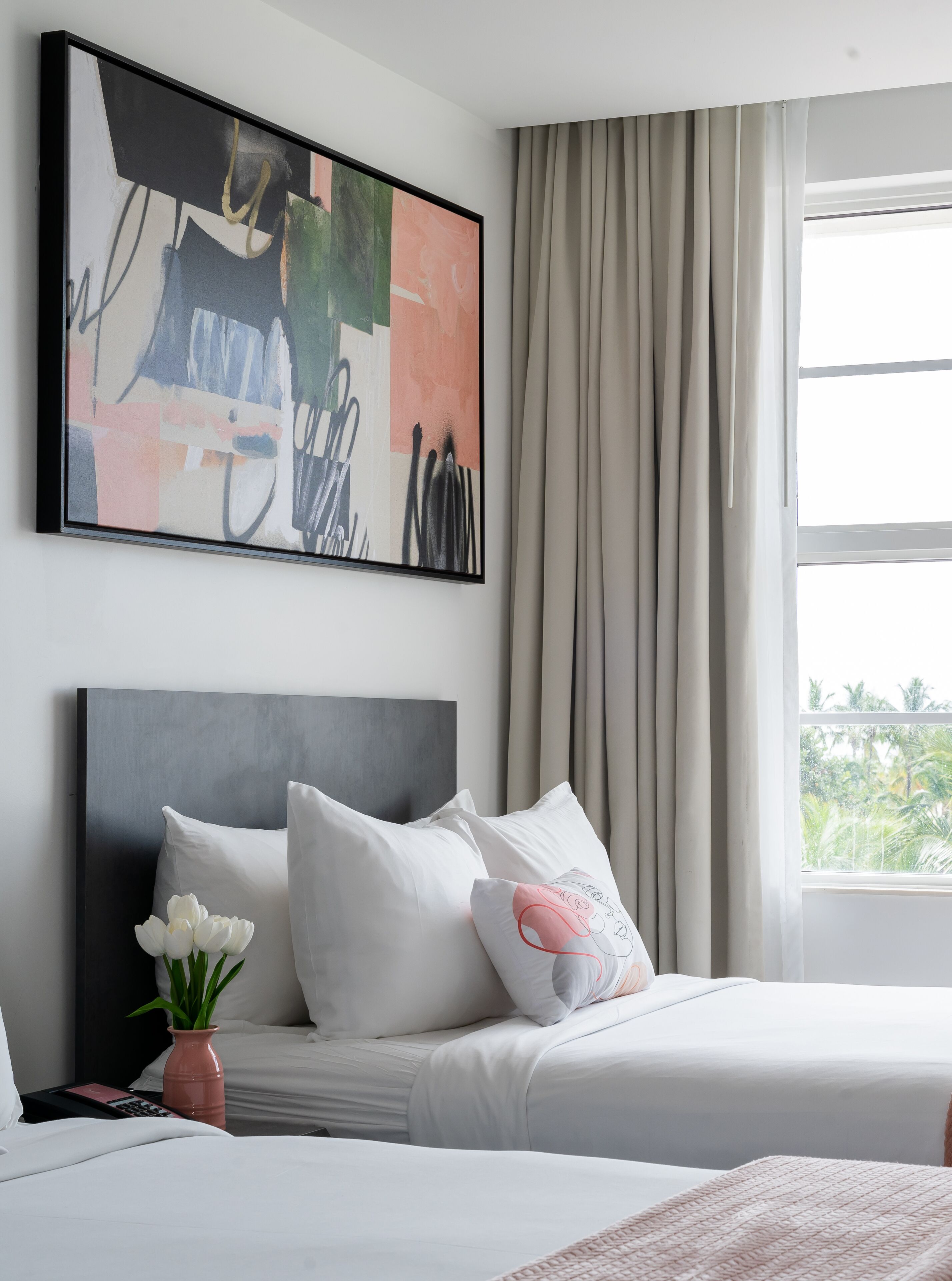 A modern hotel room with a large abstract painting, a neatly made bed with white bedding, a vase with flowers, and a view of greenery through the window.