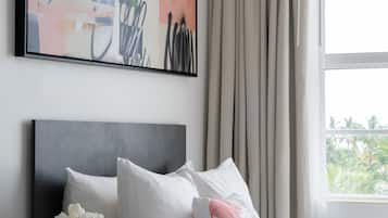 A modern hotel room with a large abstract painting, a neatly made bed with white bedding, a vase with flowers, and a view of greenery through the window.