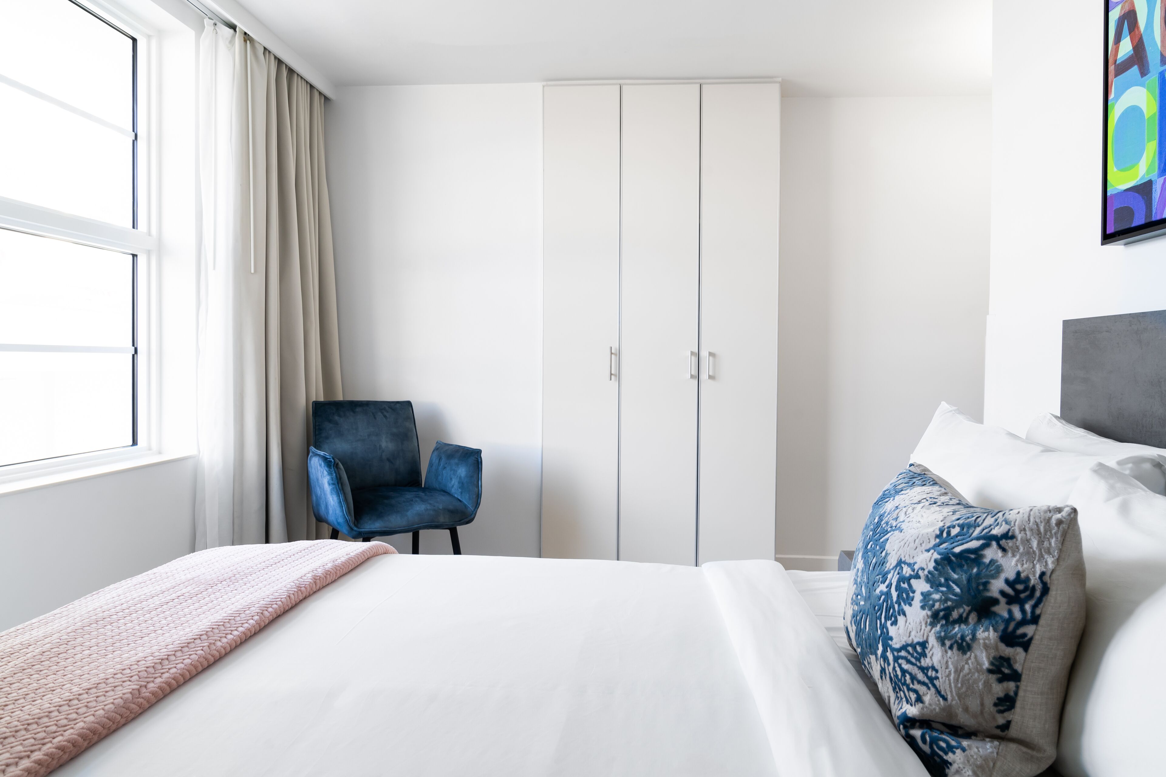 A hotel room with a bed, a chair, a wardrobe, and a painting on the wall.