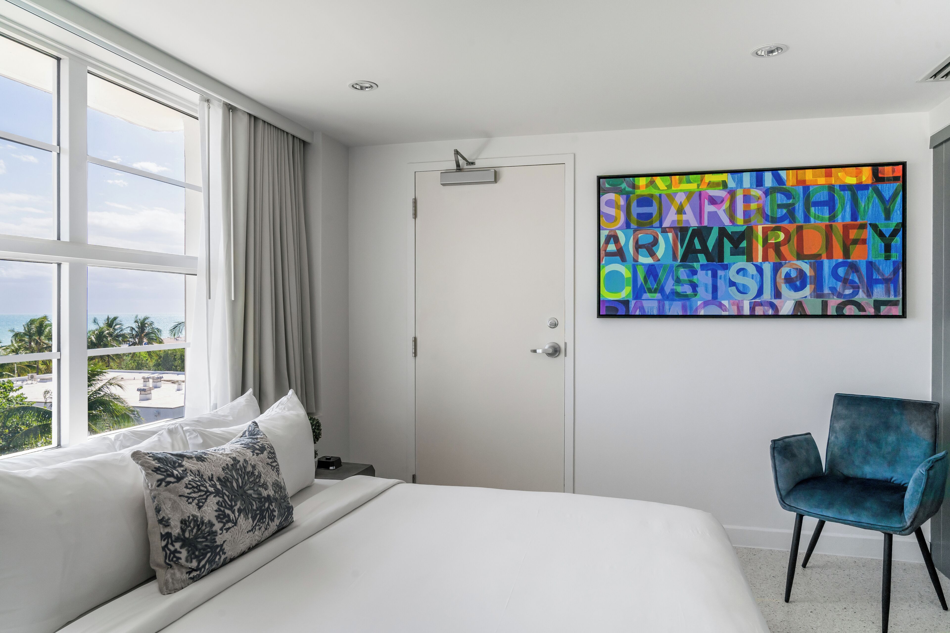 A hotel room with a large bed, a blue chair, a door, and a colorful artwork on the wall.