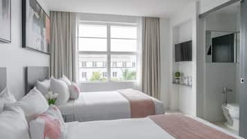 A modern hotel room with two beds, a large window with curtains, a mounted TV, and a bathroom with a toilet and sink.