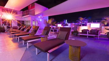A rooftop lounge area with sun loungers and a small table, surrounded by potted plants and a white building in the background.