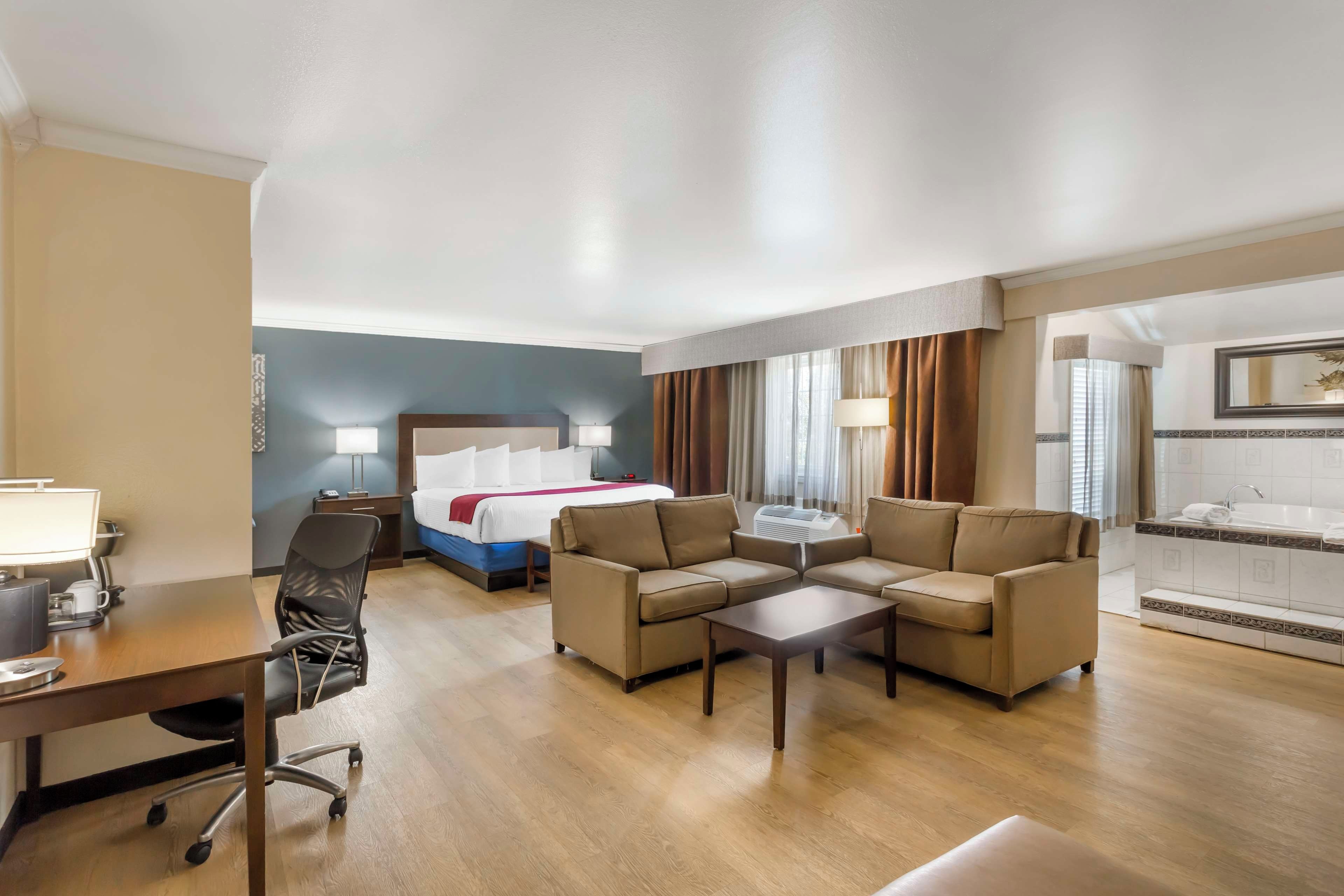 A hotel room with a bed, two sofas, a desk with a chair, a small table, and a bathroom with a bathtub.