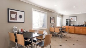 A dining area with a black table and chairs, a kitchen in the background, and framed artwork on the walls.