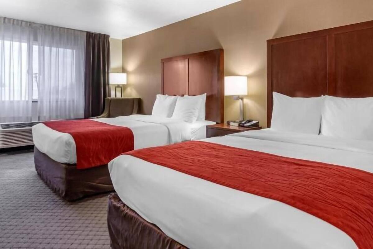 A hotel room with two beds, a large window with curtains, a wooden headboard, and a bedside table with a lamp.
