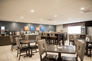 A restaurant with multiple tables and chairs, a counter with various food items, and a large screen displaying content.