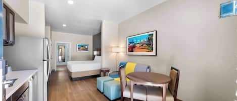 A compact hotel room with a kitchenette, a bed, a dining area with a table and chairs, a small table with a lamp, and a painting on the wall.