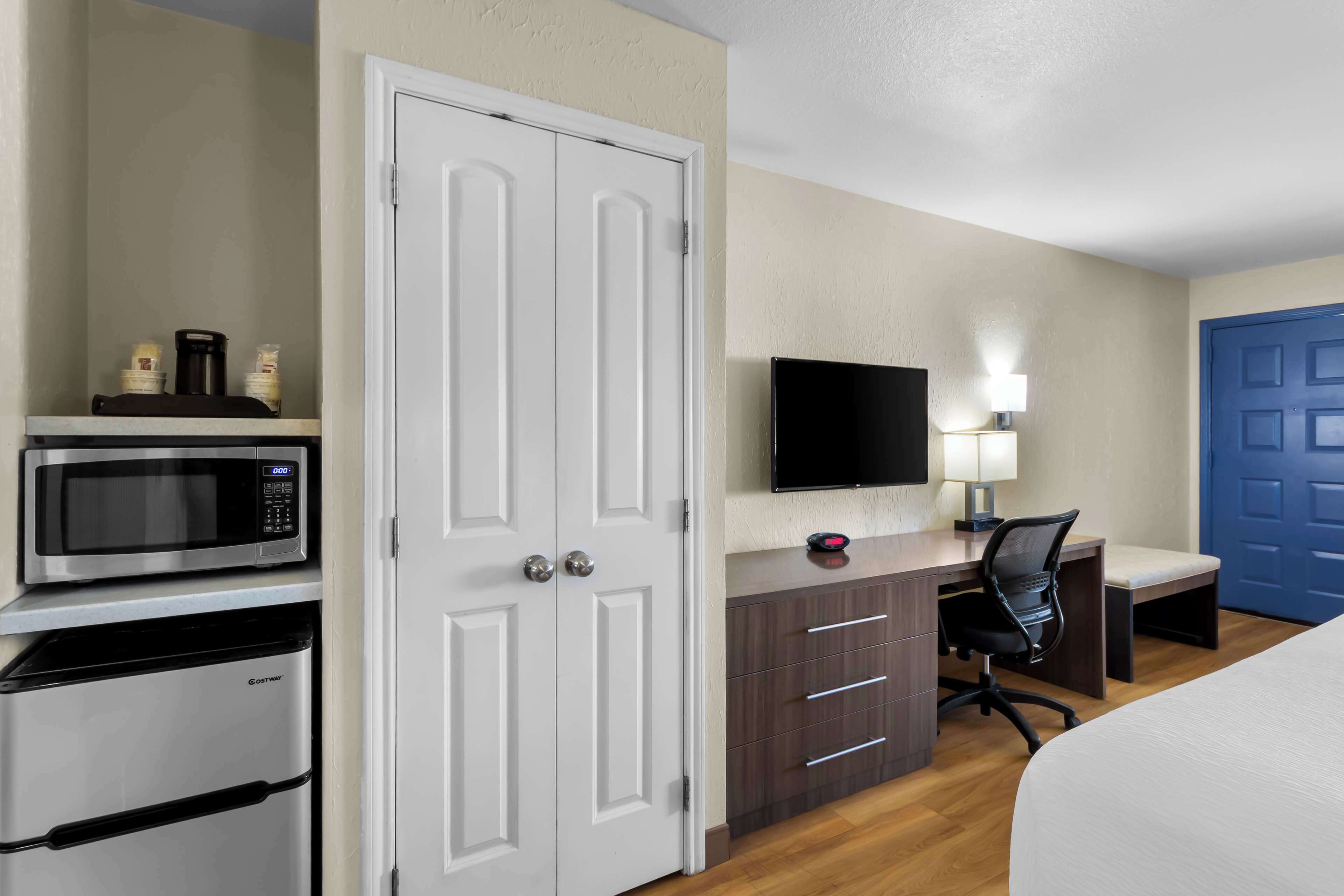 A hotel room with a white door, a flat-screen TV, a desk with a chair, a microwave, and a refrigerator.
