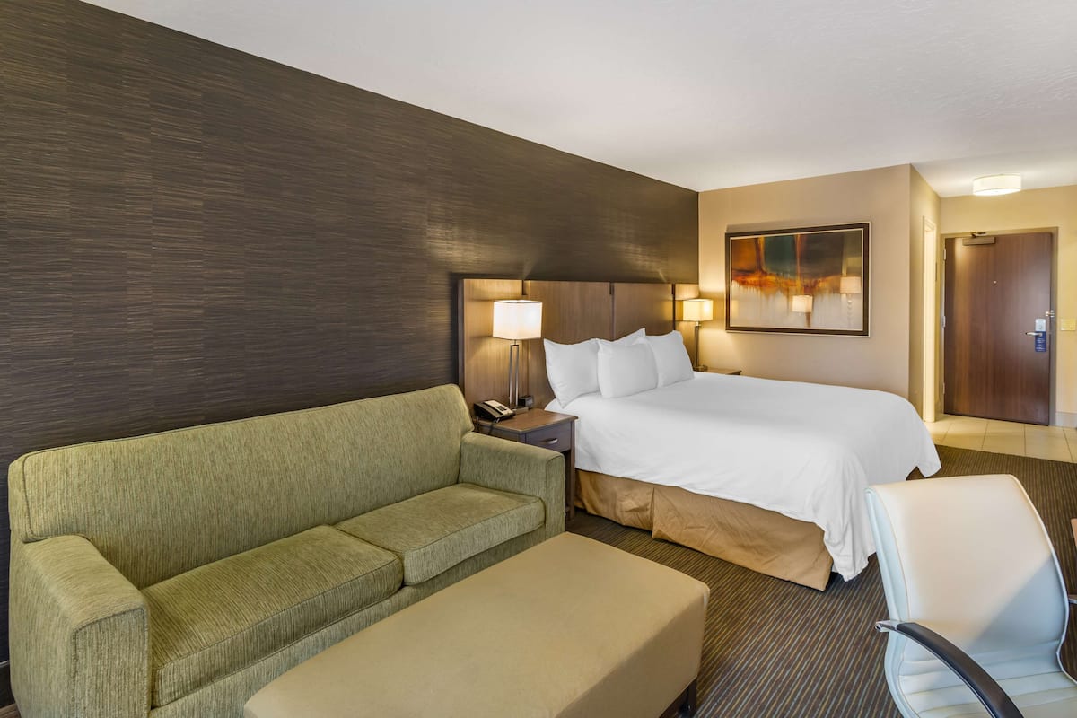 A hotel room with a bed, a green sofa, a white armchair, a nightstand with lamps, and a painting on the wall.