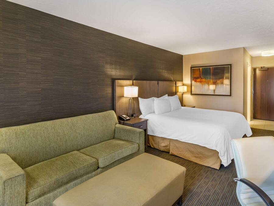 Habitación de hotel con una cama, un sofá verde, un sillón blanco, una mesita de noche con lámparas y un cuadro en la pared.
