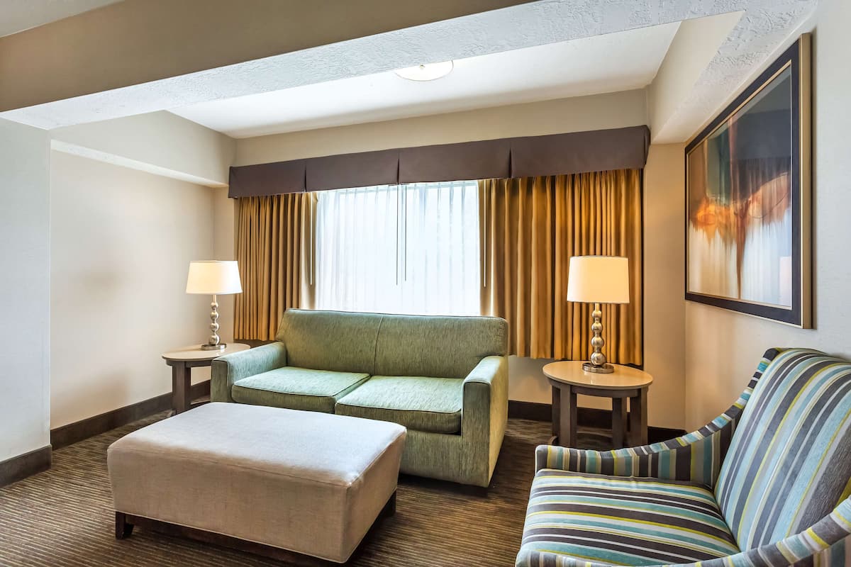 A hotel room with a green sofa, a striped armchair, and a beige ottoman. There are two lamps on side tables and a large painting on the wall.