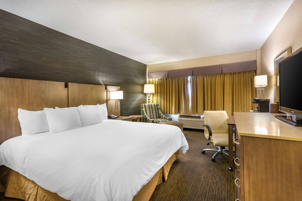 A hotel room with a large bed, a desk with a computer, a chair, a desk lamp, a TV, and a window with curtains.