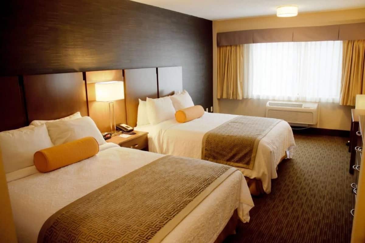 A hotel room with two beds, a bedside lamp, a telephone, a window with curtains, and an air conditioning unit.