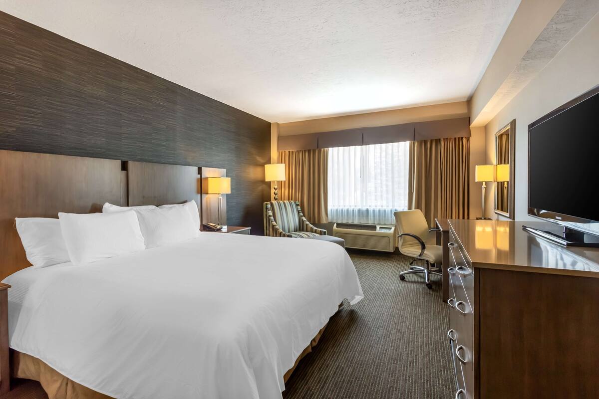 A hotel room with a large bed, a desk with a flat-screen TV, two lamps, a chair, and a large window with curtains.