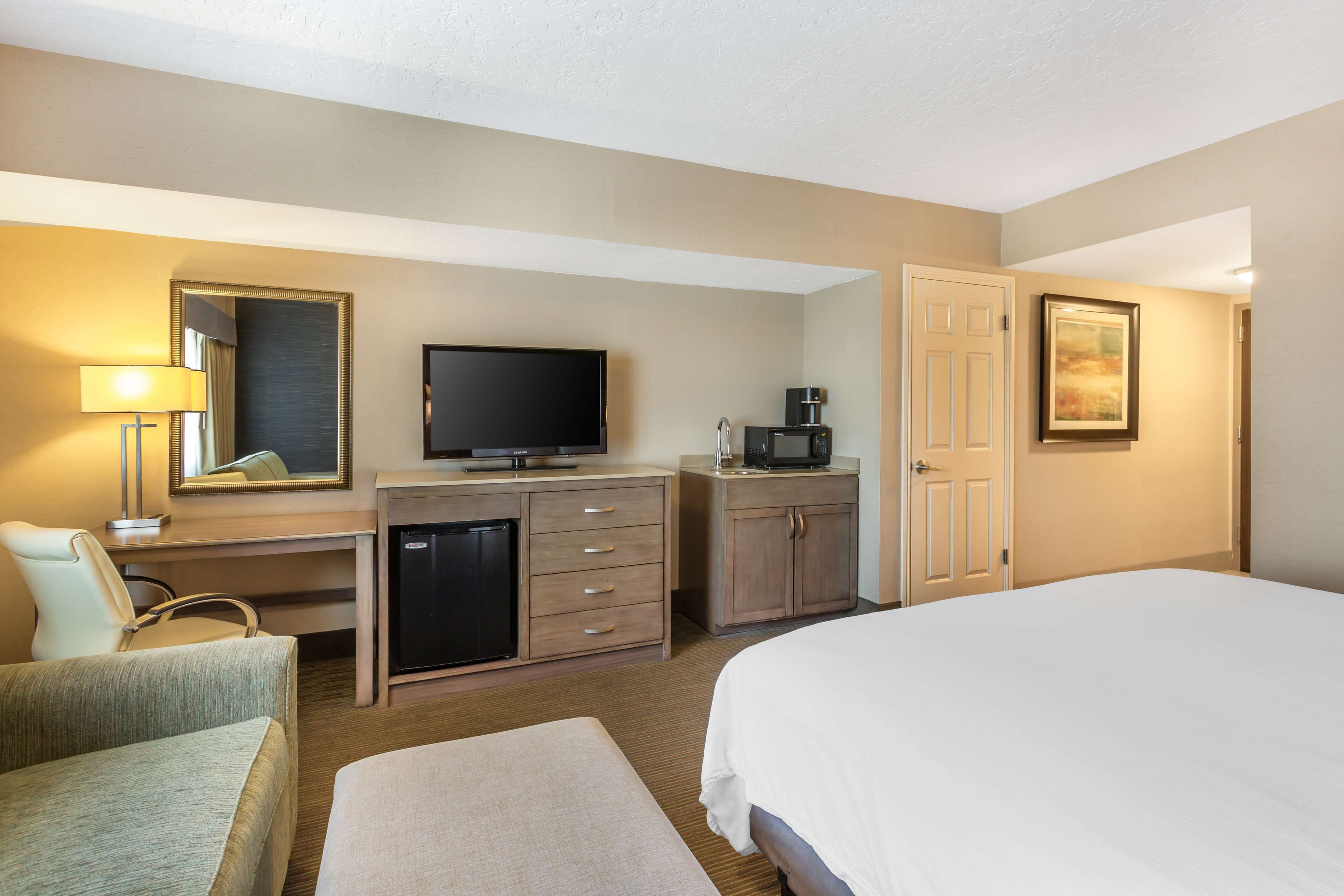 A hotel room with a bed, a desk with a television, a coffee maker, a refrigerator, a chair, and a lamp.