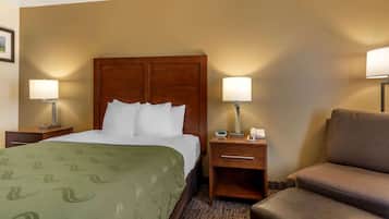 Quarto de hotel com cama de cabeceira de madeira, duas luminárias de cabeceira, colcha verde com padrão, poltrona marrom e uma pequena mesa de cabeceira com telefone e relógio.