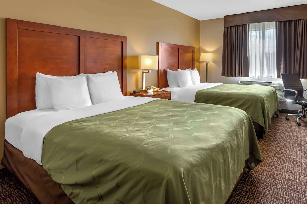 Ein Hotelzimmer mit zwei Betten, jeweils mit weißen Laken und grünen Bettdecken. Es gibt einen Schreibtisch mit einem Stuhl, eine Lampe und ein Telefon. Das Zimmer verfügt über ein großes Fenster mit Vorhängen.