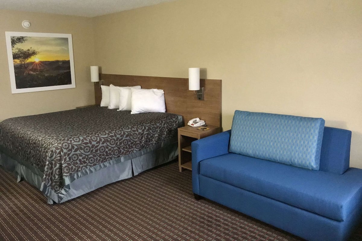 A hotel room with a bed, a blue sofa, a nightstand with a phone, and a wall-mounted lamp.