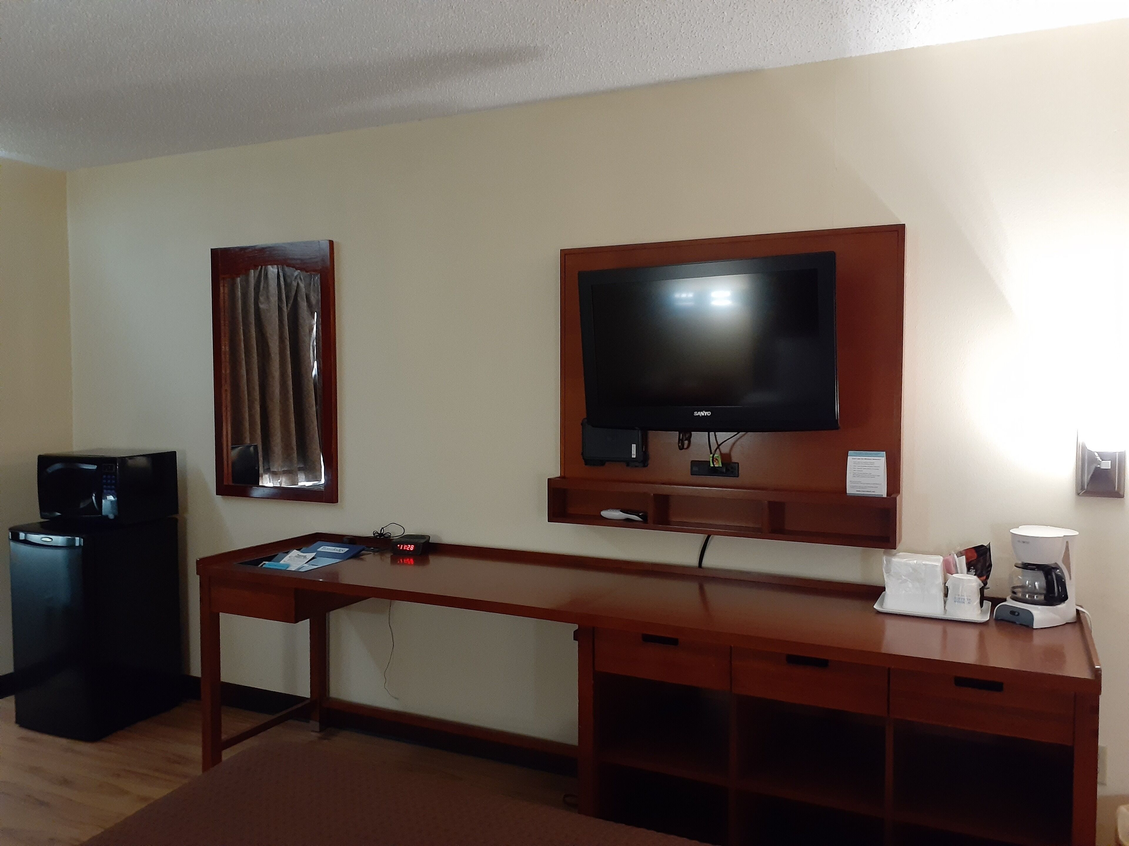 A hotel room with a wooden desk, a television mounted on the wall, a microwave, and a coffee maker.