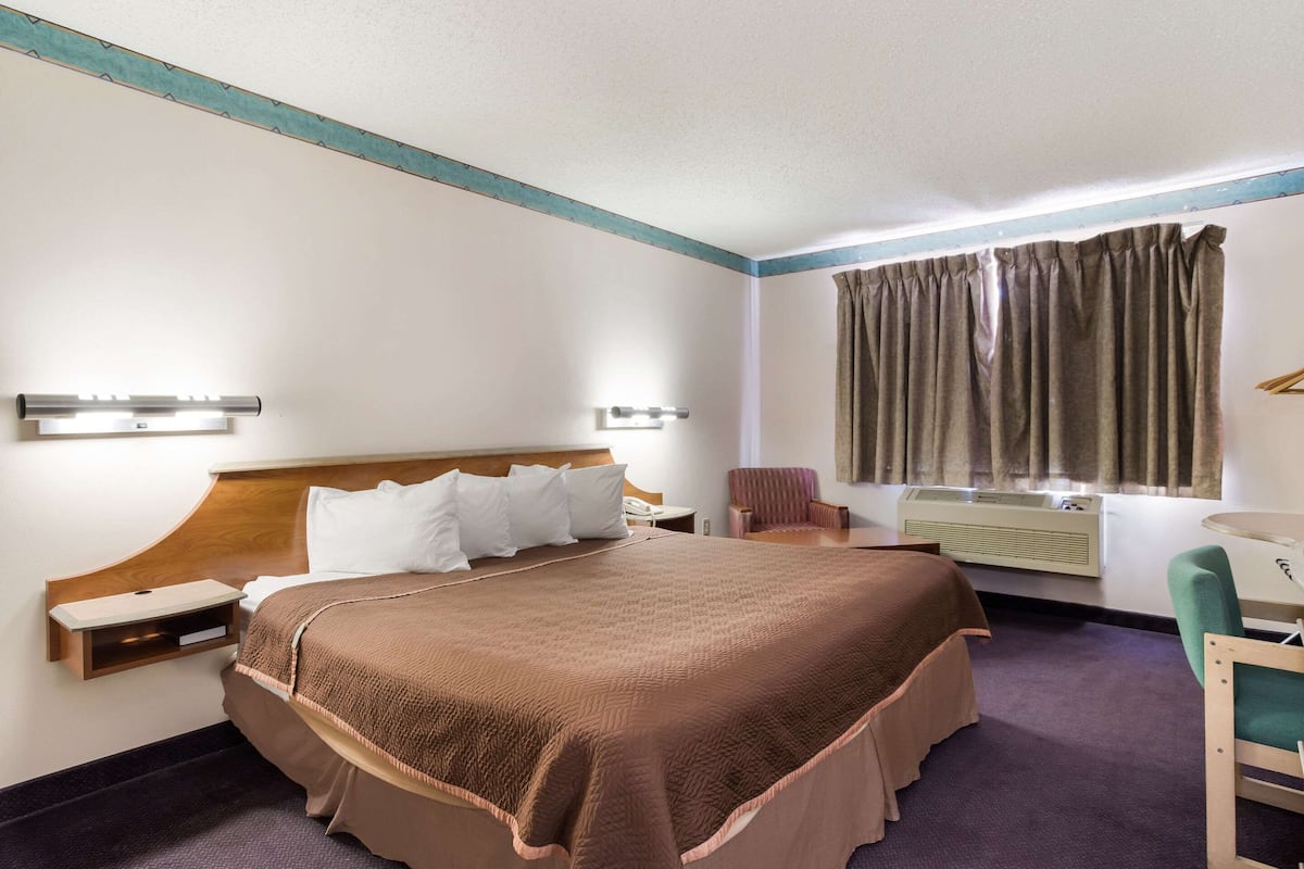 A hotel room with a large bed, bedside tables, a desk, a chair, a window with curtains, and a wall-mounted air conditioning unit.