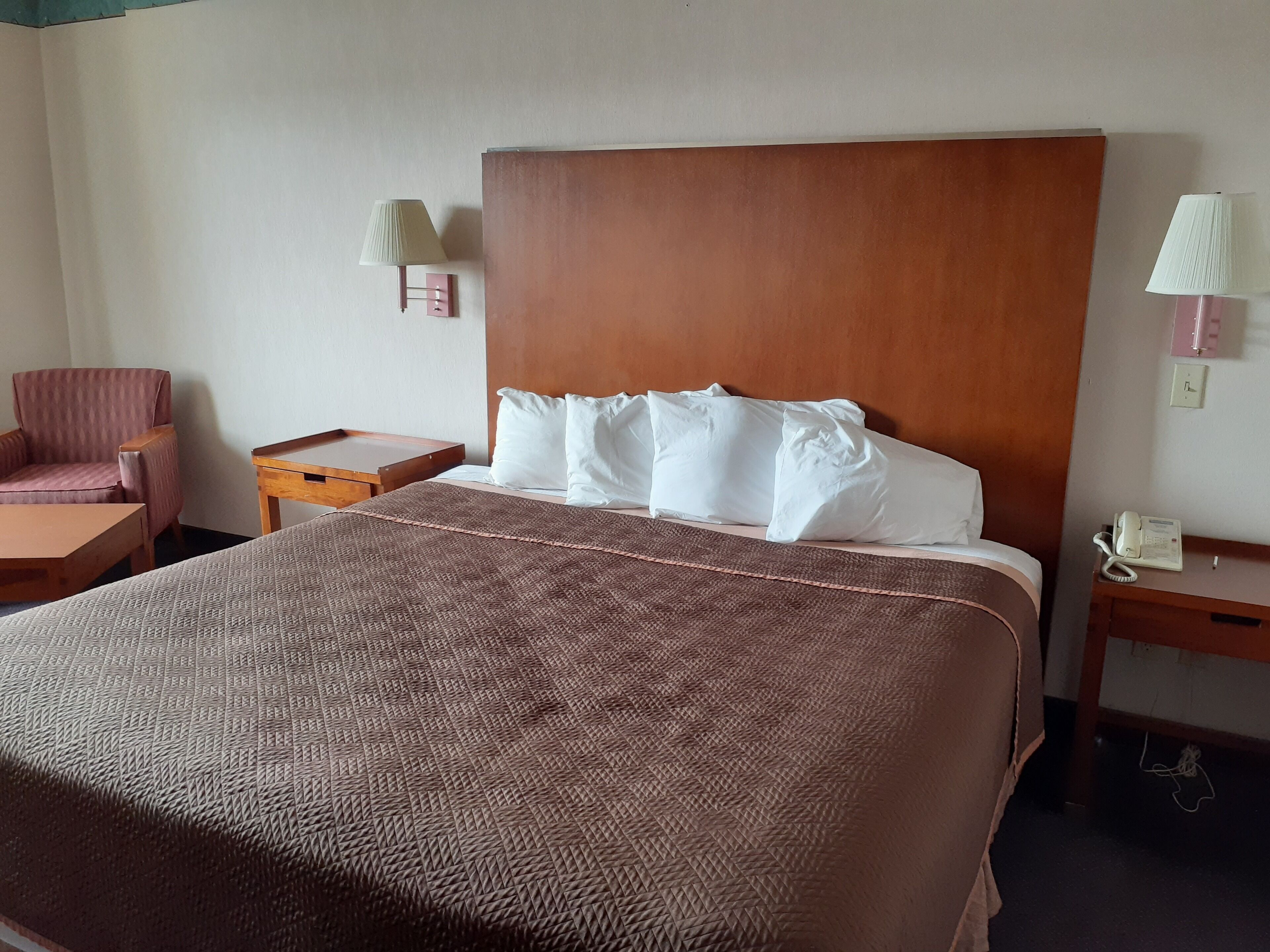 A hotel room with a large bed, a wooden headboard, a bedside table with a lamp, a chair, and a small wooden side table.