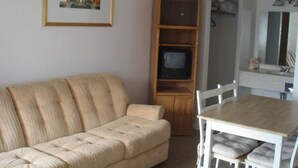 Un salon avec un canapé beige, un meuble TV en bois sur lequel est posée une télévision, une table à manger avec des chaises blanches et un tableau encadré au mur.
