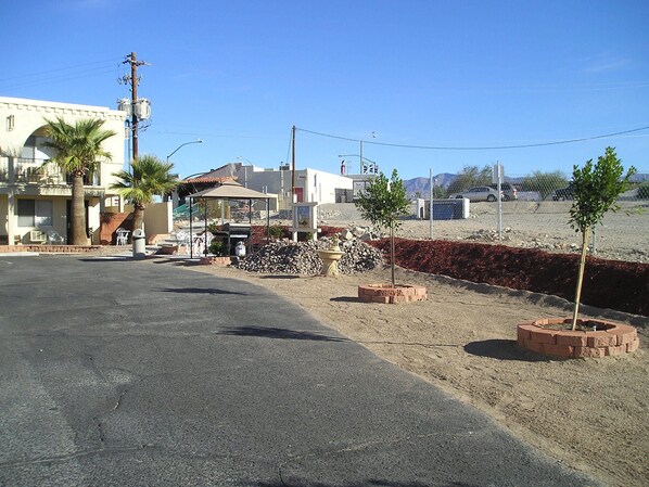 A street view with a building featuring a covered patio, palm trees, and a small garden with a tree and brick planter.