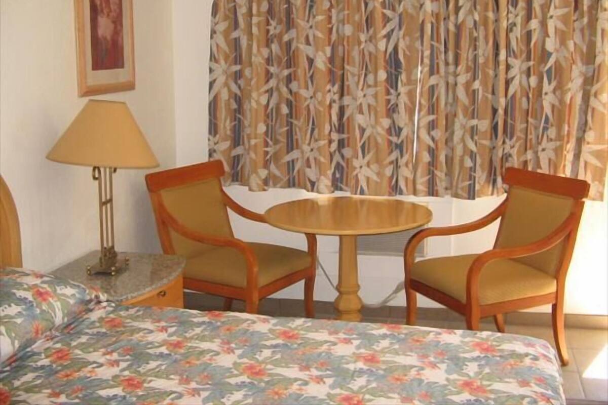 Une chambre d’hôtel avec un lit, deux chaises, une petite table, une lampe et une fenêtre avec des rideaux à motifs.