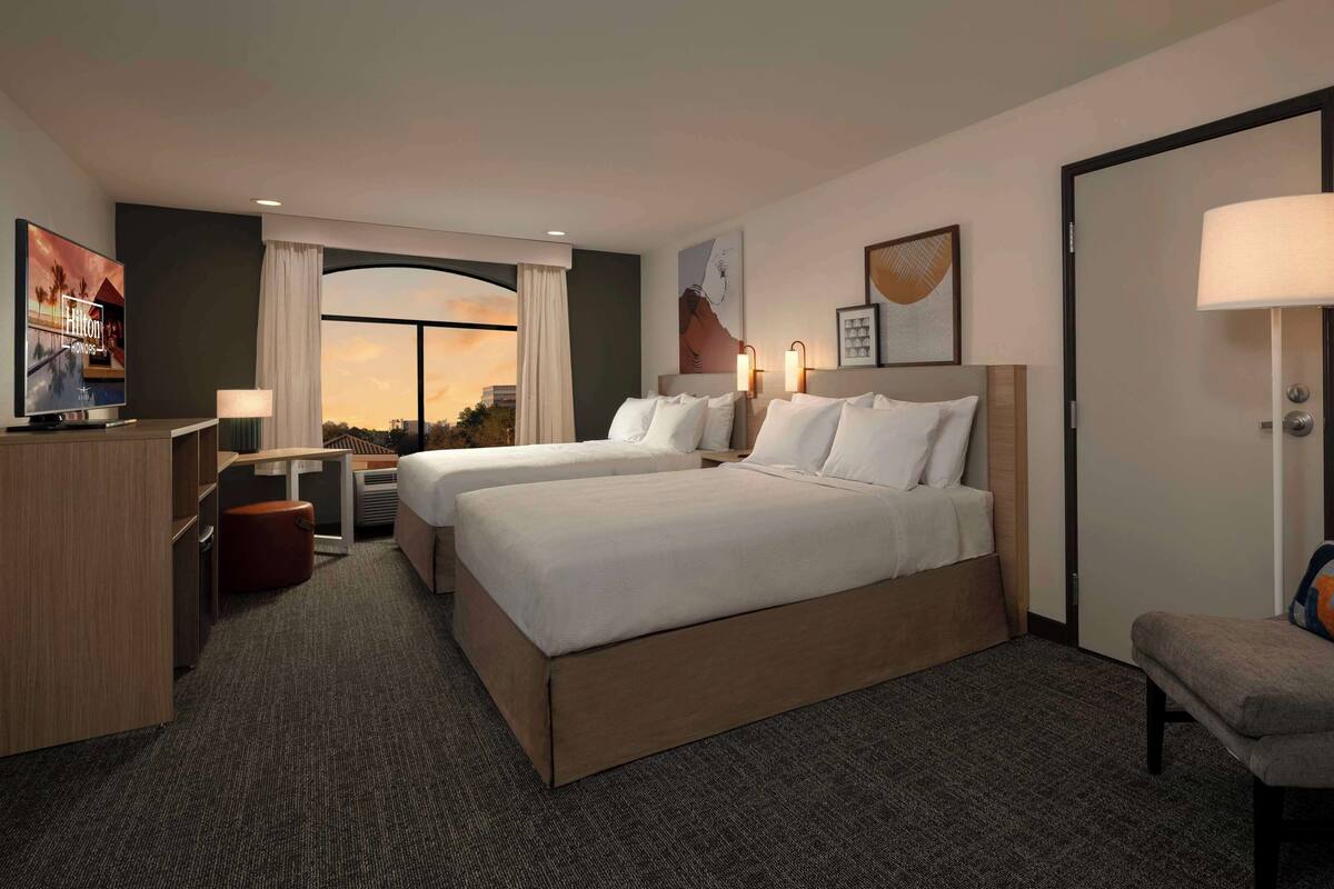 A hotel room with two beds, a desk, a chair, a TV, and a view of the sunset.