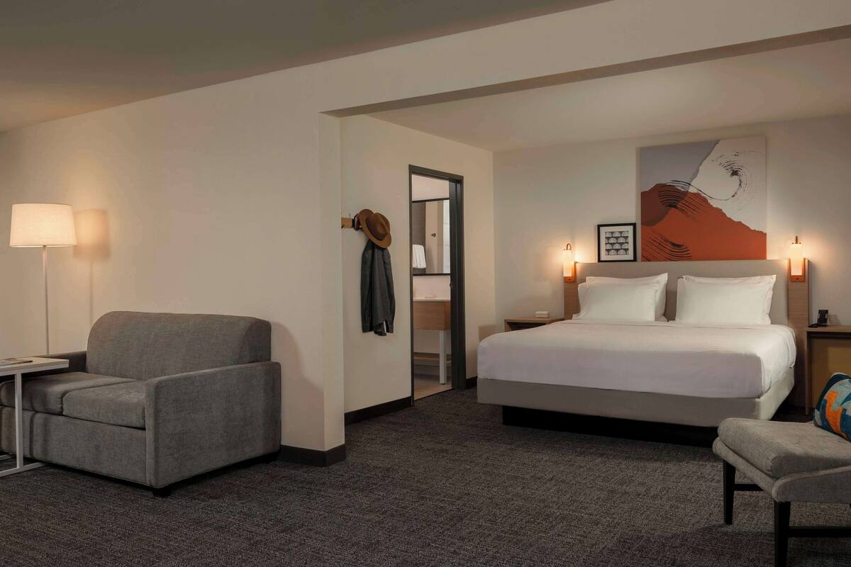 A hotel room with a large bed, two armchairs, a desk, a lamp, and a painting on the wall.