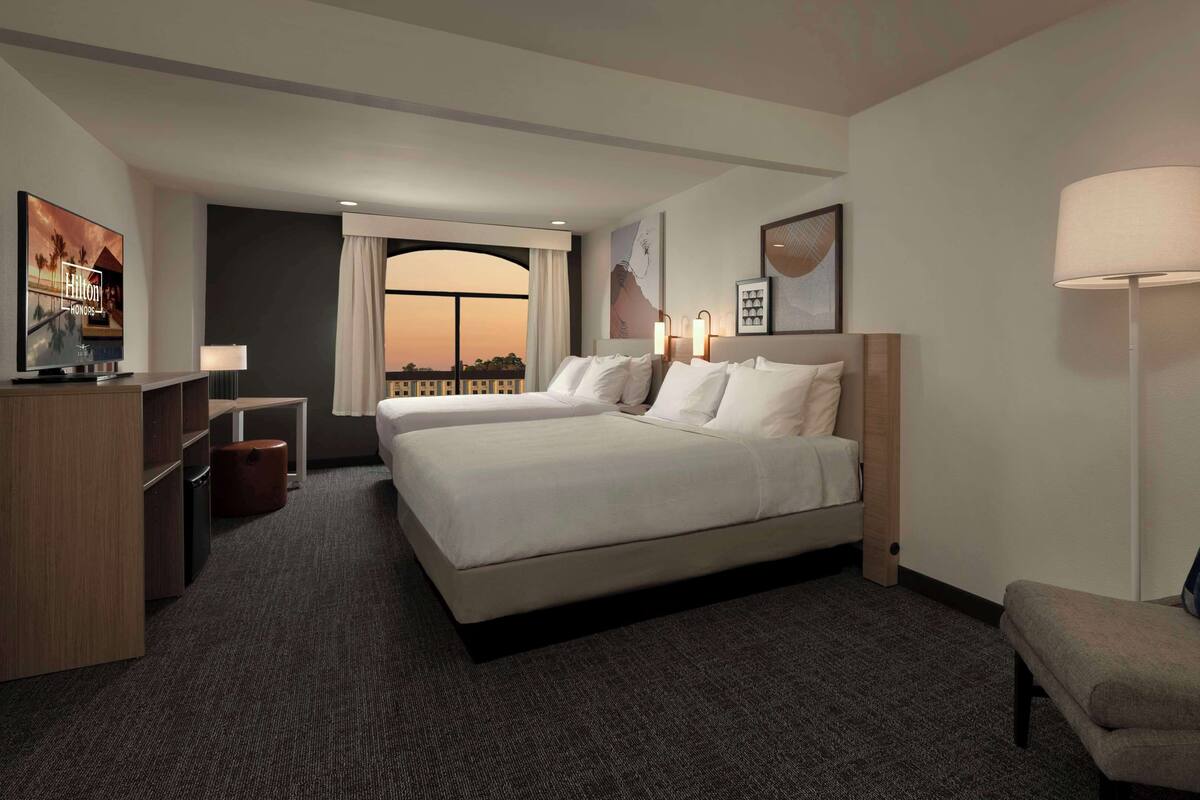 A hotel room with two beds, a desk, a chair, and a large window with a view.