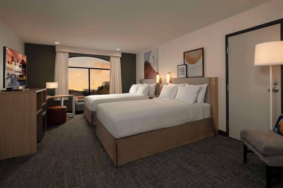 A hotel room with two beds, a desk, a chair, and a TV.